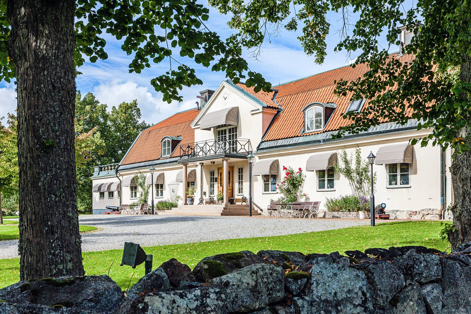 Gällfösta Herrgård framsida i solljus, tröd med gröna löv i förgrunden. Foto Martin Ekwall