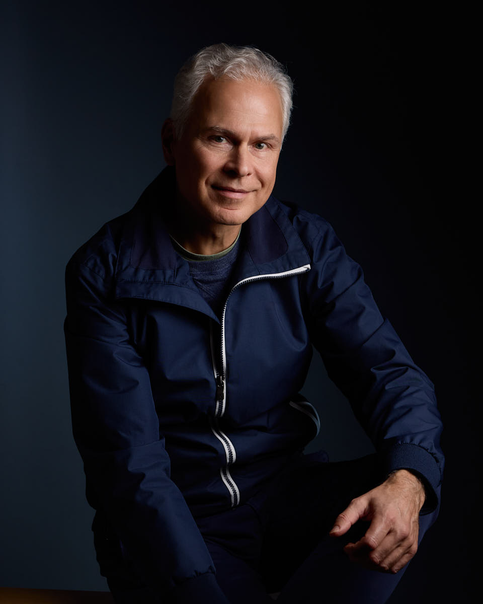 Självporträtt av Martin Ekwall, sittandes mot mörkblå bakgrund tittar in i kameran med jacka. För Photo for Brands