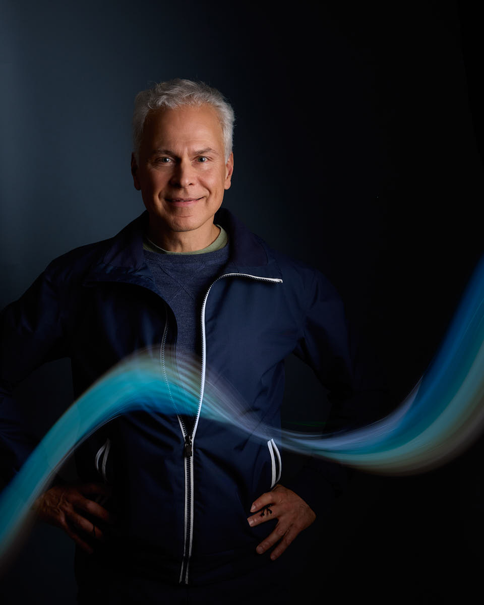 Självporträtt av Martin Ekwall, stående och leende rakt mot kameran mot mörkblå bakgrund, ljusblå lysande våg framför. För Photo for Brands